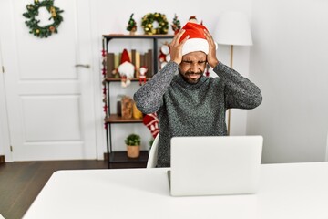 Young hispanic man with beard wearing christmas hat using laptop suffering from headache desperate and stressed because pain and migraine. hands on head.