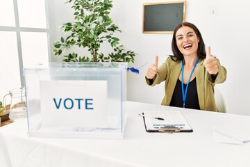 Young brunette woman sitting at election table with voting ballot approving doing positive gesture...