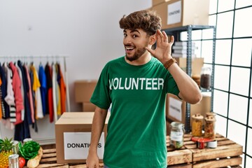 Young arab man wearing volunteer t shirt at donations stand smiling with hand over ear listening an...