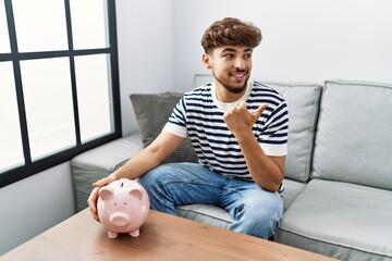Young arab man holding piggy bank pointing thumb up to the side smiling happy with open mouth