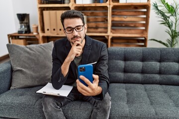 Young hispanic psychologist man doing therapy on video call with smartphone serious face thinking about question with hand on chin, thoughtful about confusing idea