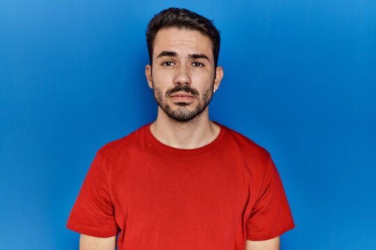 Young hispanic man with beard wearing red t shirt over blue background relaxed with serious expression on face. simple and natural looking at the camera.