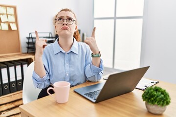 Young redhead woman working at the office using computer laptop pointing up looking sad and upset, indicating direction with fingers, unhappy and depressed.