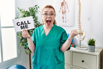 Young redhead doctor woman working at pain recovery clinic holding call me banner screaming proud, celebrating victory and success very excited with raised arms