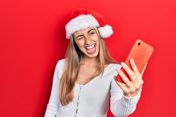 Beautiful hispanic woman wearing christmas hat using smartphone winking looking at the camera with sexy expression, cheerful and happy face.