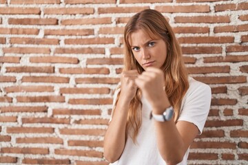 Young caucasian woman standing over bricks wall ready to fight with fist defense gesture, angry and...