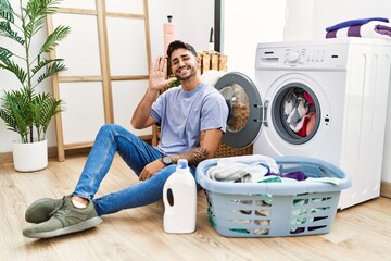 Young hispanic man putting dirty laundry into washing machine waiving saying hello happy and smiling, friendly welcome gesture