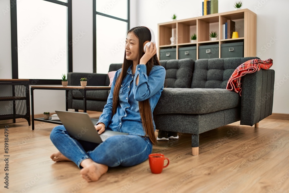 Wall mural young chinese girl using laptop and headphones sitting on the floor at home.