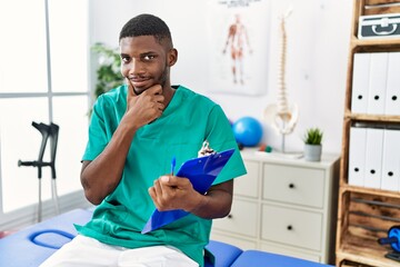 Young african american man working at pain recovery clinic looking confident at the camera smiling with crossed arms and hand raised on chin. thinking positive.