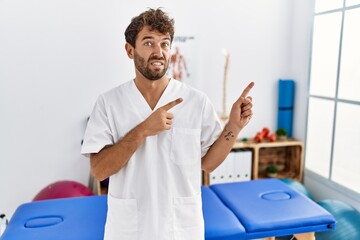 Young handsome physiotherapist man working at pain recovery clinic pointing aside worried and nervous with both hands, concerned and surprised expression