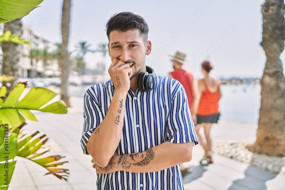 Poster Young handsome man listening to music using headphones outdoors looking stressed and nervous with hands on mouth biting nails. anxiety problem.