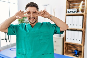 Young physiotherapist man working at pain recovery clinic doing peace symbol with fingers over face, smiling cheerful showing victory
