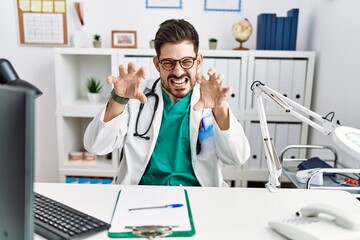 Young man with beard wearing doctor uniform and stethoscope at the clinic smiling funny doing claw gesture as cat, aggressive and sexy expression