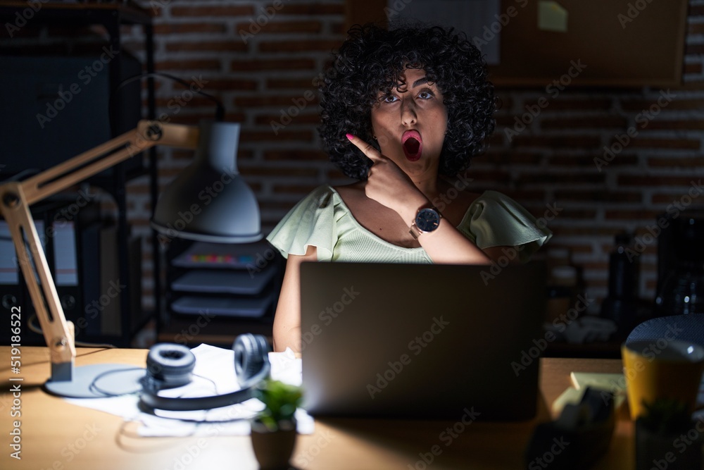 Poster Young brunette woman with curly hair working at the office at night surprised pointing with finger to the side, open mouth amazed expression.