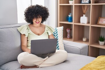 Fototapeta na wymiar Young brunette woman with curly hair using laptop sitting on the sofa at home with a happy and cool smile on face. lucky person.