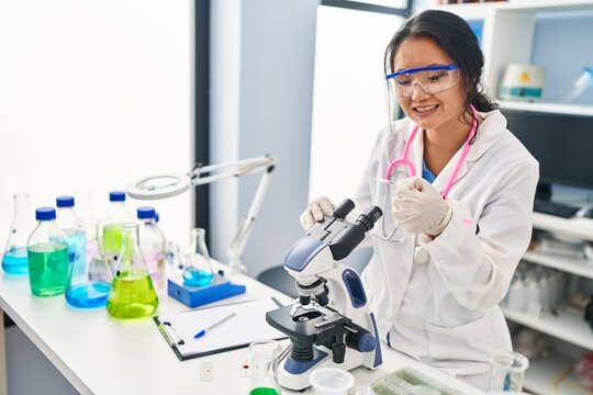 Young chinese woman wearing scientist uniform using microscope at laboratory