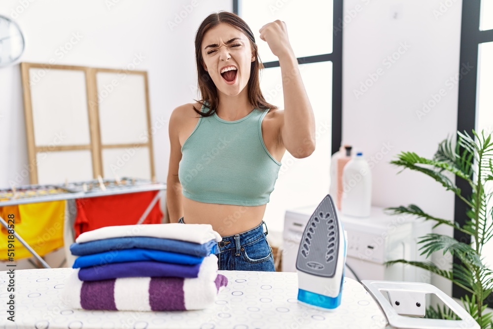 Poster Young hispanic woman ironing clothes at laundry room angry and mad raising fist frustrated and furious while shouting with anger. rage and aggressive concept.