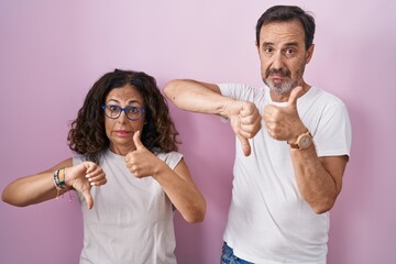 Middle age hispanic couple together over pink background doing thumbs up and down, disagreement and agreement expression. crazy conflict