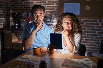 Middle age hispanic couple using touchpad sitting on the table at night thinking worried about a question, concerned and nervous with hand on chin