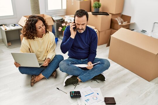 Middle age hispanic couple controlling family economy using laptop. Sitting on the floor talking on the smartphone at new home.