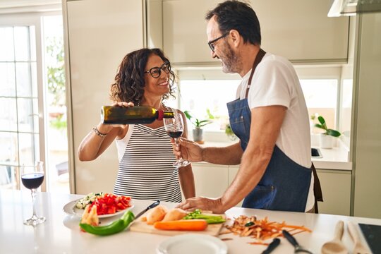 Middle age hispanic couple cooking and pouring wine on glass at kitchen