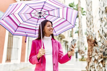 Young latin woman using smartphone holding umbrella at street