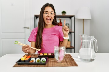Young brunette woman eating sushi using chopsticks pointing finger up with successful idea. exited and happy. number one.