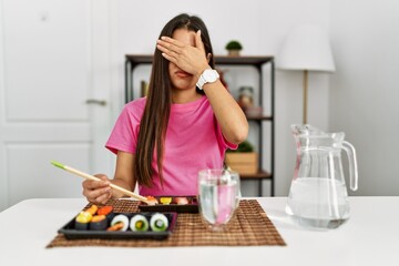 Obraz na płótnie Canvas Young brunette woman eating sushi using chopsticks covering eyes with hand, looking serious and sad. sightless, hiding and rejection concept