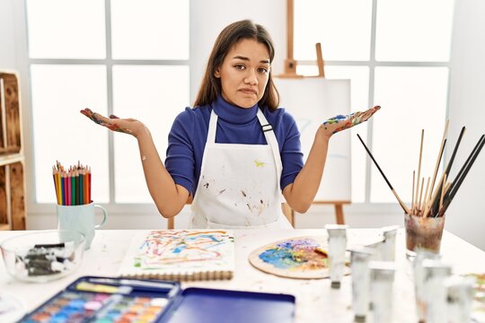 Young brunette woman at art studio with painted hands clueless and confused expression with arms and hands raised. doubt concept.