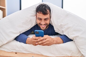 Young hispanic man using smartphone lying on bed at bedroom