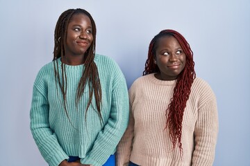 Two african woman standing over blue background smiling looking to the side and staring away...