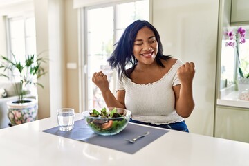 Obraz na płótnie Canvas Young hispanic woman eating healthy salad at home very happy and excited doing winner gesture with arms raised, smiling and screaming for success. celebration concept.