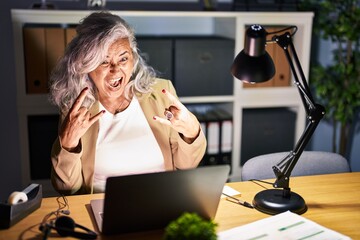 Middle age woman with grey hair working using computer laptop late at night shouting with crazy...