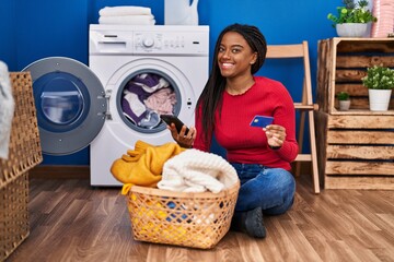 African american woman using smartphone and credit card washing clothes at laundry room