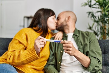 Fototapeta premium Young hispanic couple kissing and holding pregnancy test sitting on the sofa at home.