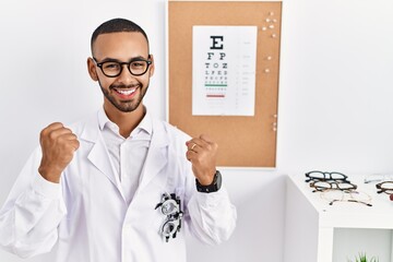 African american optician man standing by eyesight test very happy and excited doing winner gesture with arms raised, smiling and screaming for success. celebration concept.