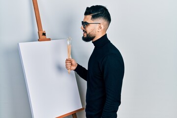 Handsome man with beard holding brushes close to easel stand looking to side, relax profile pose...