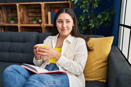 Young hispanic woman reading book and drinking coffee at home