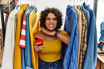 Young hispanic woman searching clothes on clothing rack using smartphone cutting throat with hand as knife, threaten aggression with furious violence