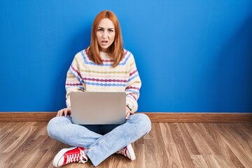 Young woman using laptop at home sitting on the floor in shock face, looking skeptical and sarcastic, surprised with open mouth