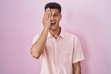 Young hispanic man standing over pink background covering one eye with hand, confident smile on face and surprise emotion.