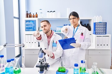 Two young people working at scientist laboratory pointing with finger to the camera and to you, confident gesture looking serious