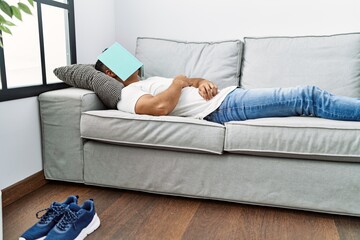 Young hispanic man lying on sofa sleeping with book on face at home