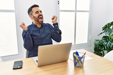Young hispanic man with beard working at the office with laptop celebrating surprised and amazed for success with arms raised and eyes closed. winner concept.