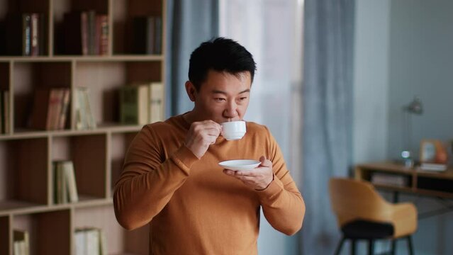 Tranquil middle aged asian man enjoying aromatic espresso, smelling cup of fresh coffee, resting at home, free space
