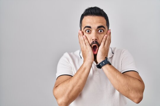 Young hispanic man with beard wearing casual clothes over white background afraid and shocked, surprise and amazed expression with hands on face