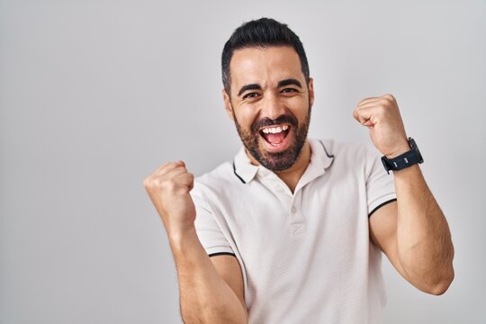 Young hispanic man with beard wearing casual clothes over white background celebrating surprised and amazed for success with arms raised and eyes closed. winner concept.