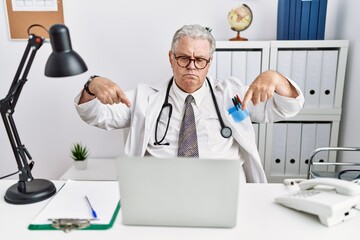 Senior caucasian man wearing doctor uniform and stethoscope at the clinic pointing down looking sad and upset, indicating direction with fingers, unhappy and depressed.