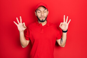 Hispanic man with beard wearing delivery uniform and cap relaxed and smiling with eyes closed doing meditation gesture with fingers. yoga concept.