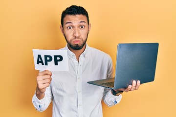 Hispanic man with beard holding computer laptop and app banner puffing cheeks with funny face. mouth inflated with air, catching air.